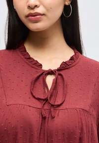 Γυναίκα που φοράει υφασμάτινο βυσσινί μπλουζάκι με βολάν στον λαιμό και δεμένο φιόγκο, φορεί επίσης λεπτό κρίκο σκουλαρίκι.