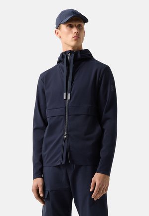 Mężczyzna ubrany w granatową bluzę z kapturem i zamkiem błyskawicznym, dopasowane spodnie z boczną kieszenią oraz granatową czapkę, stojący na jednolitym jasnym tle.