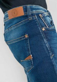 Jean en denim bleu avec une patch en cuir marron sur la taille, des garnitures en métal argenté, et des détails de couture distincts, présentant un design de poche latérale.