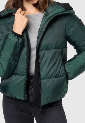 Femme portant une doudoune vert foncé sur une chemise grise et un jean noir, une main tenant le col de la veste et l'autre dans la poche.