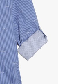 Blauw en lichtblauw gestreept overhemd met lange mouwen en opgerolde manchet, bedrukt met de woorden "ATELIER," "SCOTCH," en "MISTER."