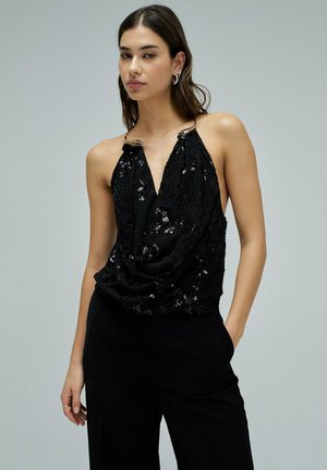 Jeune femme portant un haut noir sans manches orné de sequins avec un col en V profond et un pantalon noir, debout devant un fond clair uni.