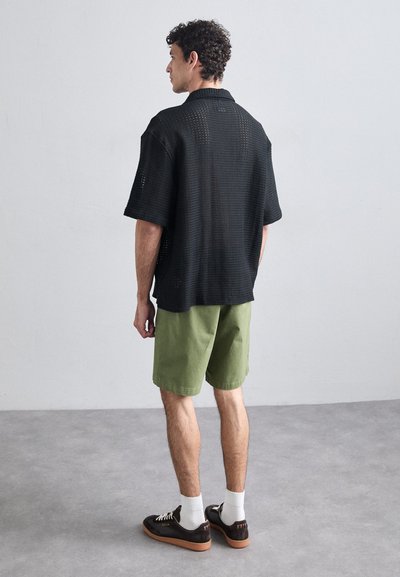 Chemise noire à mailles ouvertes à manches courtes, shorts verts, et chaussures marron avec des chaussettes blanches ; présente une coupe décontractée et un design informel.