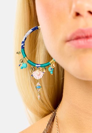 Creolen mit grünen und blauen, gewebten Akzenten, goldfarbenem Beschlag und dekorativen Anhängern, darunter eine Muschel, Perlen und ein tropfenförmiger Edelstein.