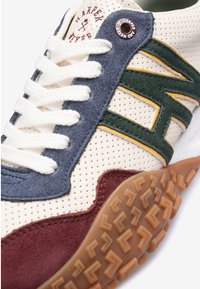 Zapatilla con parte superior de cuero crema perforado, acentos de ante en azul, verde y burdeos, cordones blancos y suela de goma texturizada en marrón.