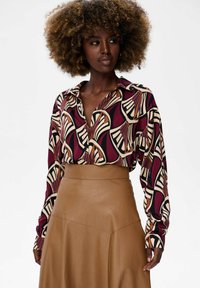 Woman with curly hair wearing a patterned long-sleeve blouse tucked into a high-waisted tan leather skirt, looking to the side.