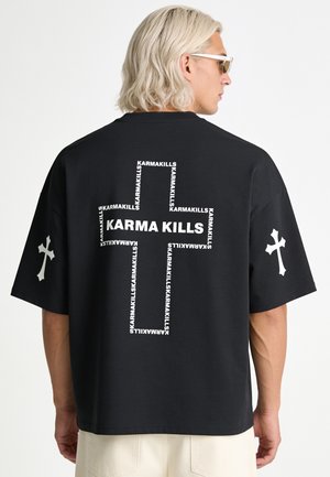 Person mit hellblonden Haaren, die eine Sonnenbrille trägt und ein schwarzes T-Shirt mit weißem Text, der ein Kreuz bildet, sowie Kreuzsymbole auf den Ärmeln.