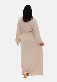 Robe longue beige avec des manches longues, taille froncée, tissu fluide et texture lisse, portée avec des chaussures à talons clairs.