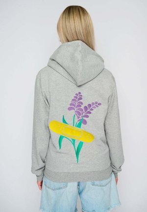 Donna con lunghi capelli biondi indossa una felpa grigia con cappuccio con fiori viola e una grande grafica di cerotto giallo sulla schiena, abbinata a pantaloncini di jeans chiari.