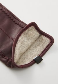 Burgunderfargede læderhandsker med en blød, beige uldforing. Synlige stingdetaljer langs kanterne og et lille sort mærke på siden.
