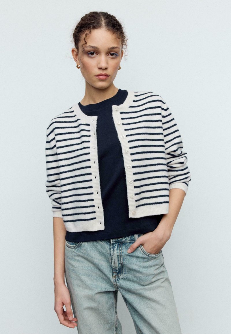 Cardigan en maille rayée crème et marine, avec fermetures à boutons et poignets côtelés. Porté sur un haut foncé avec un jean coupe décontractée.