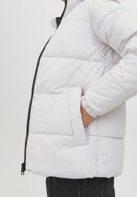 Biała puchowa kurtka z poziomym pikowaniem, czarnym zamkiem błyskawicznym i boczną kieszenią, z elastycznymi mankietami i wysokim kołnierzem.