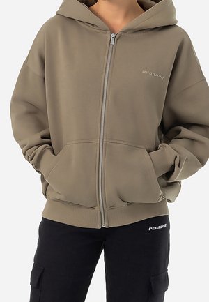 Person trägt einen braunen Kapuzenpullover mit Reißverschluss und Fronttaschen sowie eine schwarze Hose mit Cargotaschen, beide mit der Marke "Pegador".