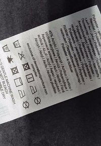 Vårdtag etikett av vävd tyg som visar skötselråd på flera språk, med ikoner för tvätt och torkning. Vit bakgrund, svart text.