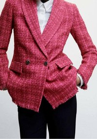 Vaaleanpunainen tweed-kaksoisnappinen bleiseri, jossa on teksturoitu pinta ja fringe-yksityiskohtia helman kohdalla, mustia nappeja. Päällä valkoinen paita.