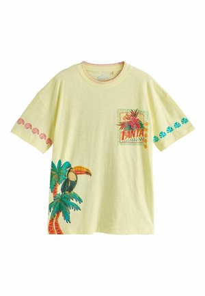 Tânără femeie stând pe nisip, purtând un tricou galben supradimensionat cu imprimeu tropical, ținând o geantă mică asortată, papuci turcoaz și brățări.