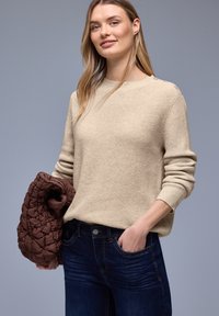 Femme portant un pull en maille beige et un jean bleu foncé, tenant une veste matelassée marron pliée devant un fond gris.