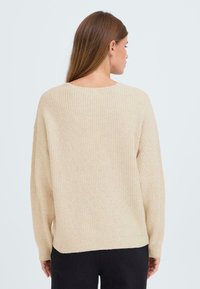 Pull en tricot beige au fit décontracté, texture côtelée, manches larges et col rond. Le dos est uni avec un ourlet légèrement en baisse.