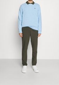 Ljusblå sweatshirt med grön krage, kombinerad med mörkgröna byxor och vita sneakers. Lacoste-logotyp på sweatshirten. Enkelt mönster.