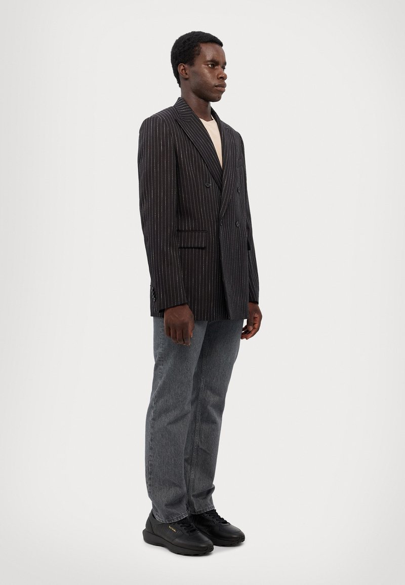 Dobbeltradet sort blazer med pinstriber og klapkrave, parret med grå jeans og sorte sneakers, som har en afslappet pasform og minimale detaljer.