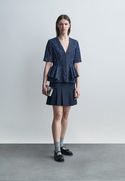 Femme debout portant un haut bleu texturé à basque avec des boutons, une jupe plissée marine, des chaussettes grises, des mocassins noirs, et tenant une petite pochette.