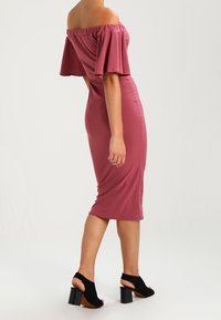 Robe midi rose, décolleté bardot, manches évasées, silhouette ajustée et tissu lisse ; associée à des sandales à talons noires.