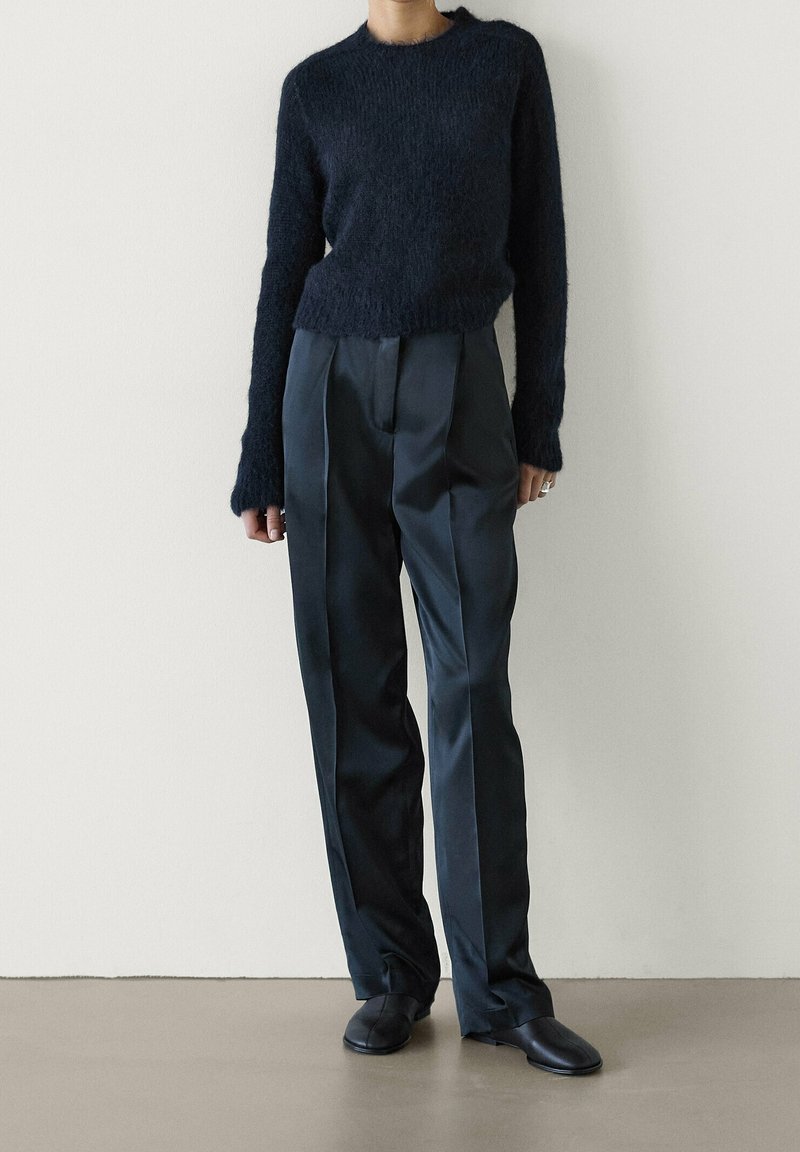 Personne portant un pull noir duveteux à manches longues, un pantalon noir plissé taille haute et des chaussures noires devant un mur clair uni.