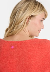 Strickpullover in leuchtendem Korallenrot, mit einem weiten Ausschnitt und einem kleinen rosa Knopfdetail auf der Rückseite. Weicher Stoff mit gemütlicher Passform.