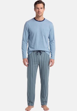 Uomo in piedi a piedi nudi che indossa una camicia azzurro chiaro a maniche lunghe e pantaloni del pigiama a righe blu su uno sfondo uniforme.