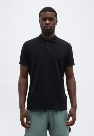 Tume-nahaline mees, kes kannab musta lühikeste varrukatega polotrikooga ja rohelist lühikest pükst, seisab lihtsa valge tausta ees.