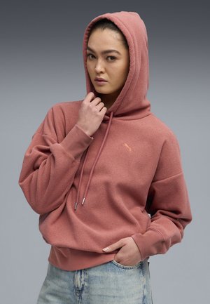 Hoodie rosa con tessuto testurizzato, con corde di regolazione e un piccolo logo. Presenta una vestibilità comoda e polsini a costine, abbinato a jeans chiari.