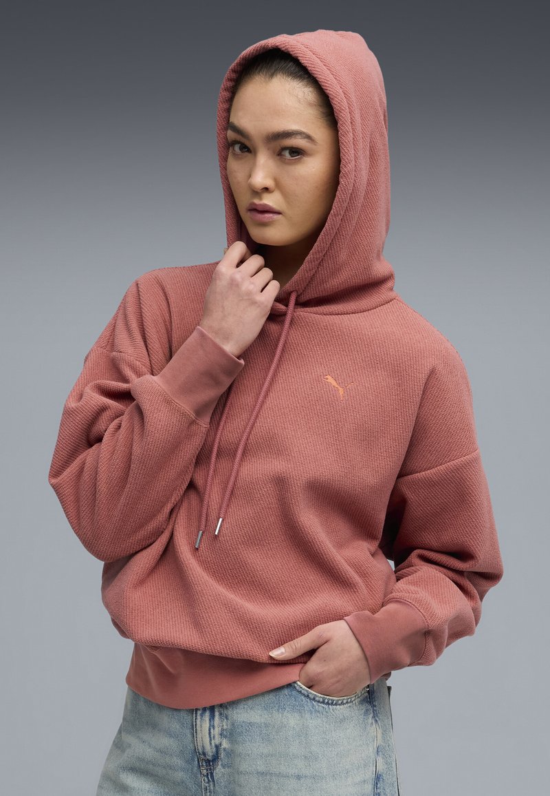 Hoodie rosa con tessuto testurizzato, con corde di regolazione e un piccolo logo. Presenta una vestibilità comoda e polsini a costine, abbinato a jeans chiari.