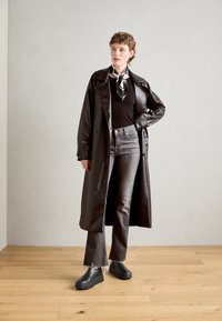 Long black trench coat with glossy finish, paired with a black top, gray flared jeans, and black chunky shoes. Scarf features a black-and-white pattern.