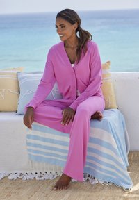 Roza kardigan in usklajene hlače iz lahkega materiala, sedeča na svetli dekici z modrimi črtami, ob vzglavnikih in z morskim razgledom.