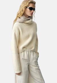 Maglione a coste color crema con collo a imbuto, abbinato a pantaloni larghi con vita elastica di un leggero color beige. Completano l'outfit occhiali da sole scuri e una sciarpa morbida.