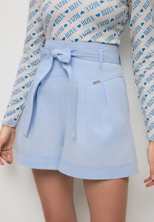 Jasnoniebieskie szorty z wysokim stanem z przednimi zakładkami i wiązanym paskiem, zestawione z koszulą z długim rękawem ozdobioną wzorem powtarzającego się napisu "BIZZU" w kolorze niebieskim.