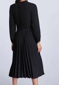 Robe noire plissée avec des manches longues, un col volanté et une ceinture en tissu à la taille, avec des plis verticaux le long du dos.