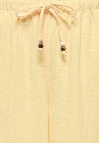 Taille à cordon sur tissu jaune clair avec deux cordons terminés par des perles marron et blanches nouées en nœud.