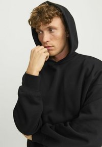 Black hooded sweatshirt with ribbed cuffs, relaxed fit, and a soft texture. Individual is gently grasping their chin.