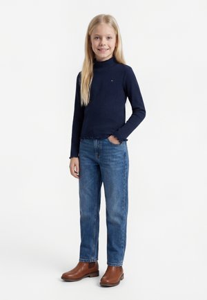 Ung jente står, iført en marineblå ribbet langermet topp, blå jeans og brune ankelstøvler, smilende mot kameraet.