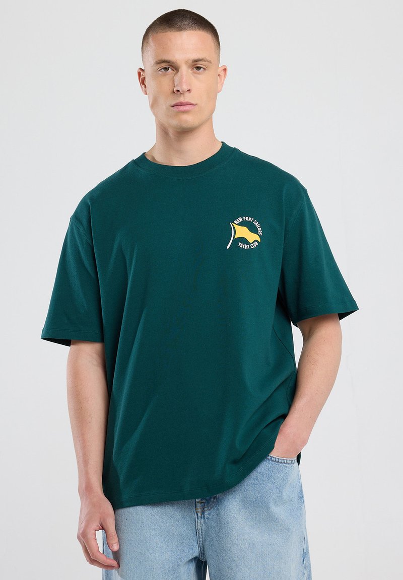 Vihreä puuvillapaita lyhyillä hihoilla, pyöreällä kaula-aukolla ja keltainen lippu kuvio painettuna vasemmassa rinnassa.