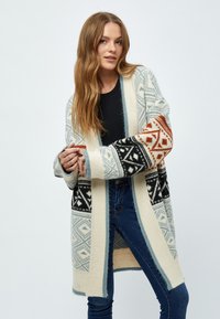 Cardigan long en tricot présentant des motifs géométriques en crème, noir, orange et bleu. Devant ouvert avec une coupe décontractée et des manches larges.