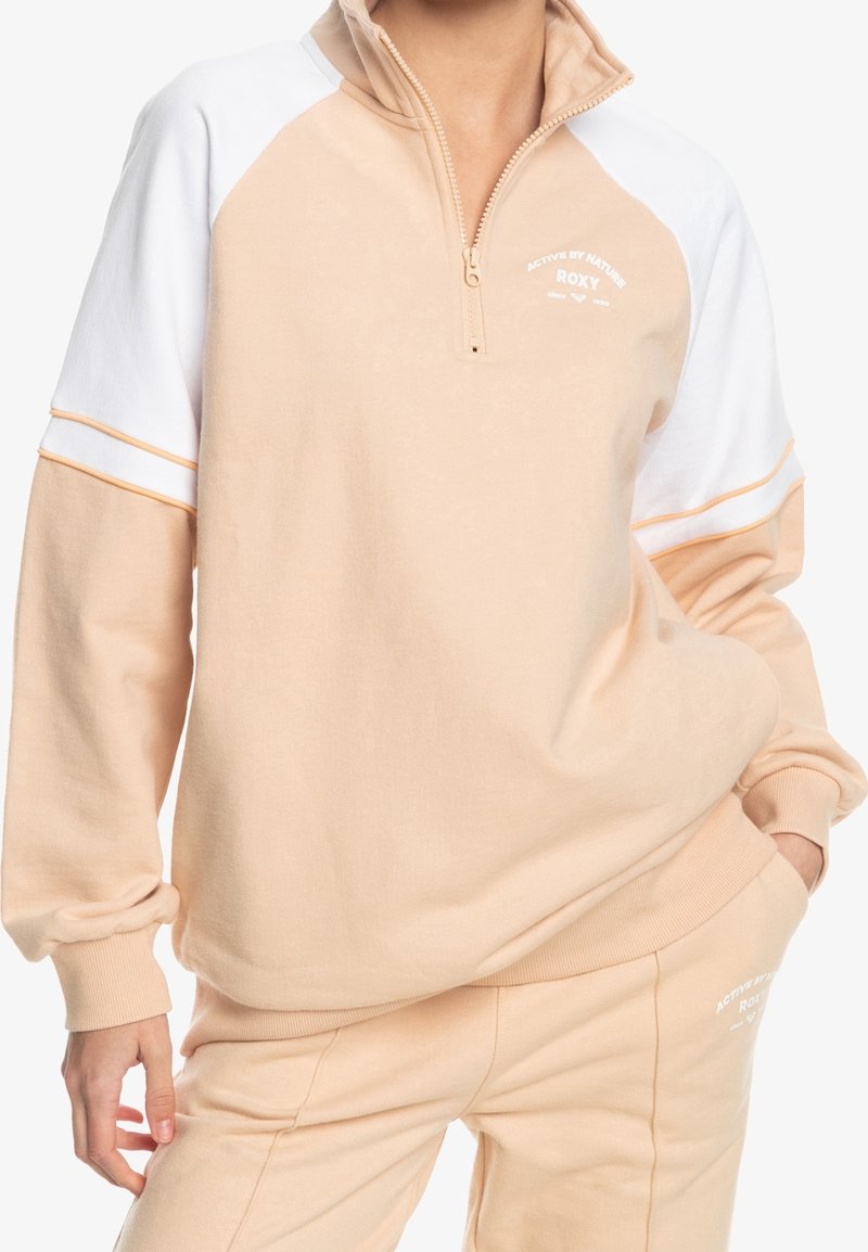 Personne portant un sweat-shirt à fermeture éclair quart beige et blanc et un pantalon beige assorti avec le logo "Roxy", debout avec une main dans la poche.