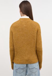 Senapsgul stickad tröja med en strukturig yta, med rund hals och ribbad nederkant, ihop med ljusblå jeans.