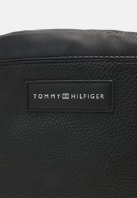 Borsa nera in pelle testurizzata con cuciture a vista. Presenta un'etichetta rettangolare nera con testo in rilievo bianco: "TOMMY HILFIGER."