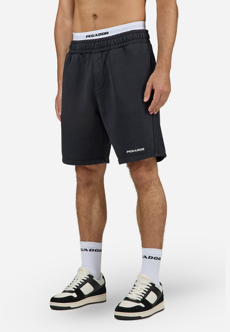 Pantaloni corti neri in cotone con cintura elastica, caratterizzati da un logo bianco "PEGADOR" e tasche laterali. Abbinati a scarpe da ginnastica bianche e nere.