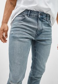 Vaqueros de mezclilla azul claro con un corte ajustado, que cuentan con una rodilla desgastada, cinco bolsillos y un cierre de botón en la cintura.