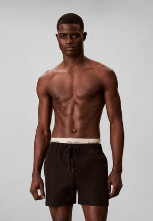 Oberkörperfreier Mann, der schwarze Badeshorts mit Kordelzug und sichtbarem weißen Calvin Klein-Bund trägt, vor schlichtem grauem Hintergrund stehend.