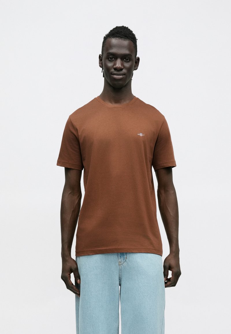 Brun bomuld T-shirt med rund halsudskæring og korte ærmer. Har en lille grafisk logo på brystet. Glat tekstur, afslappet pasform.