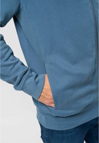 Blue sweatshirt with a zipper, featuring a front pocket, ribbed cuffs, and soft texture. The design includes a fitted silhouette.